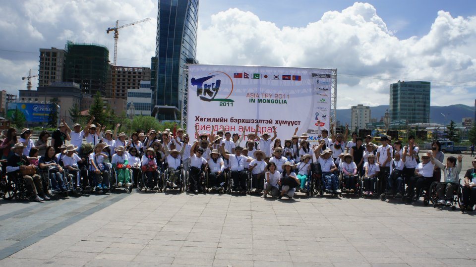 Улаанбаатарын төв талбайд «Asia TRY 2011 in Mongolia» самбарын өмнө тэргэнцэртэй болон зогсож буй олон оролцогч гараа өргөн хамтдаа зургаа авахуулж байна.