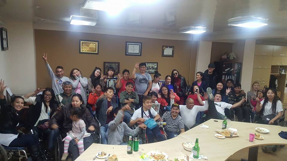 Танхимд ширээний ард хамт олон гараа өргөн баяр тэмдэглэж байна; тэргэнцэртэй оролцогчид урд эгнээнд.